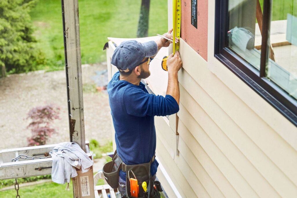Construction worker using a tape measure to mark beige siding on a house.