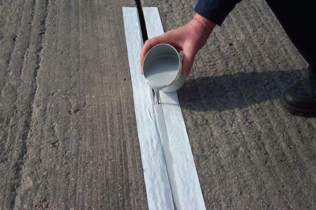 A person pours a liquid sealant from a small container into a crack in concrete pavement.