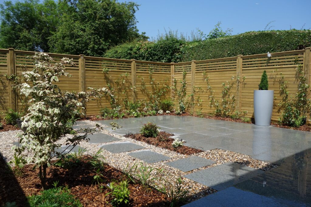 Modern garden patio featuring grey stone paving, stepping stones, gravel borders, and a high wooden slatted fence.