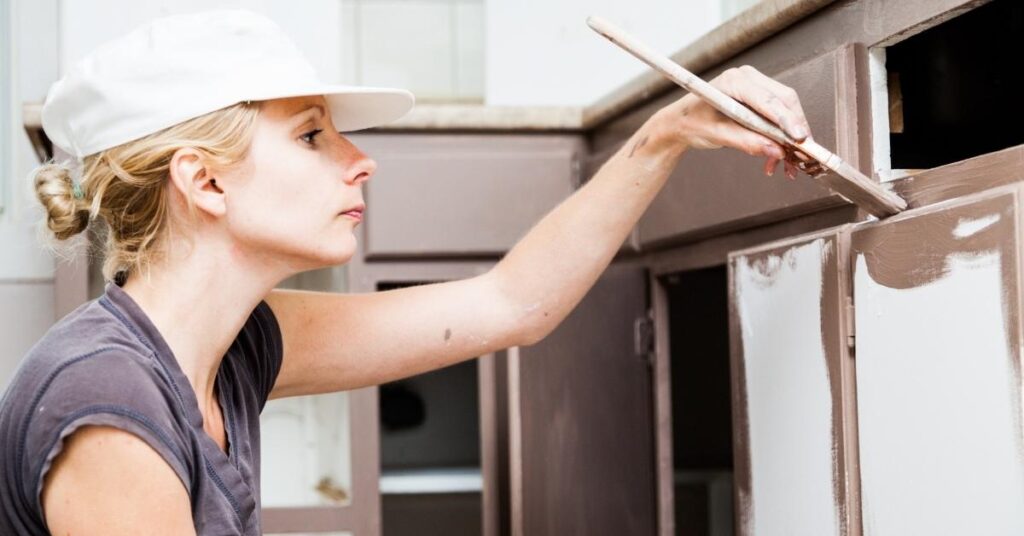 A woman in a white cap uses a paintbrush to apply brown paint to kitchen cabinets.