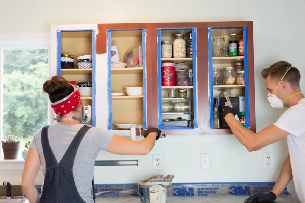 Two people painting open kitchen cabinets reddish-brown, using blue painter's tape and protective gear.