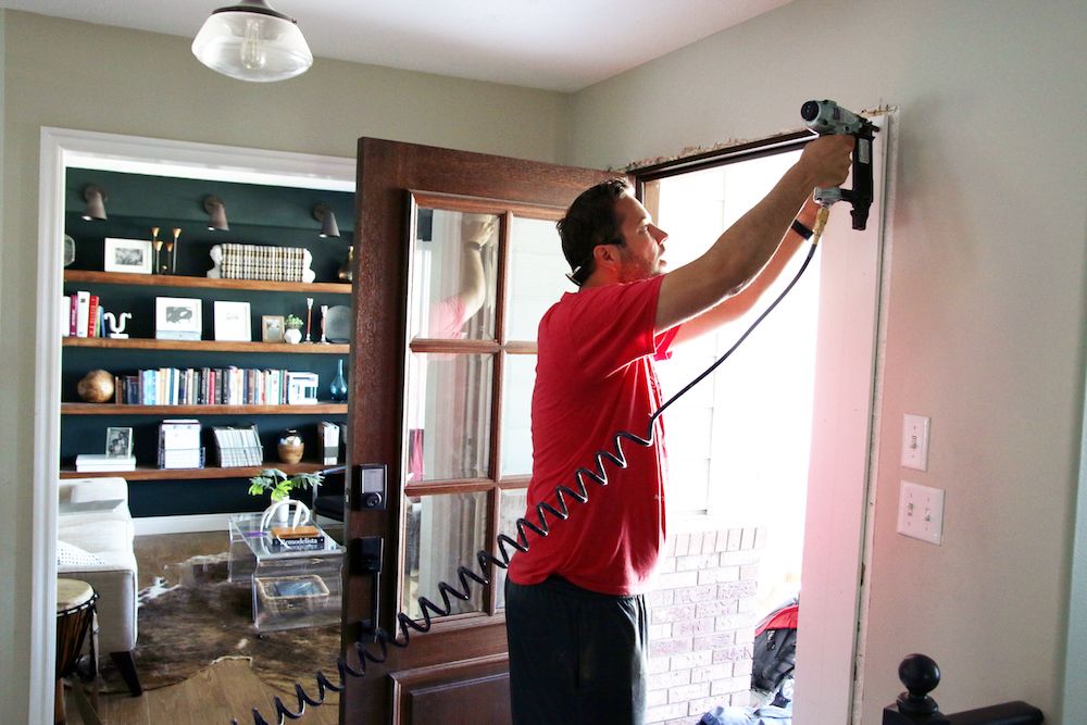 Man in a red shirt installing a wooden door frame with a pneumatic nail gun.