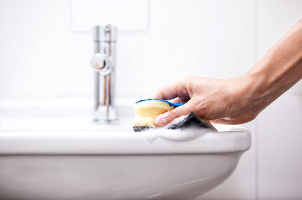 A hand cleaning the edge of a white bathroom sink with a soapy sponge.