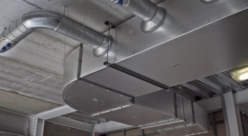 Industrial silver HVAC ducts and piping mounted to a concrete ceiling.