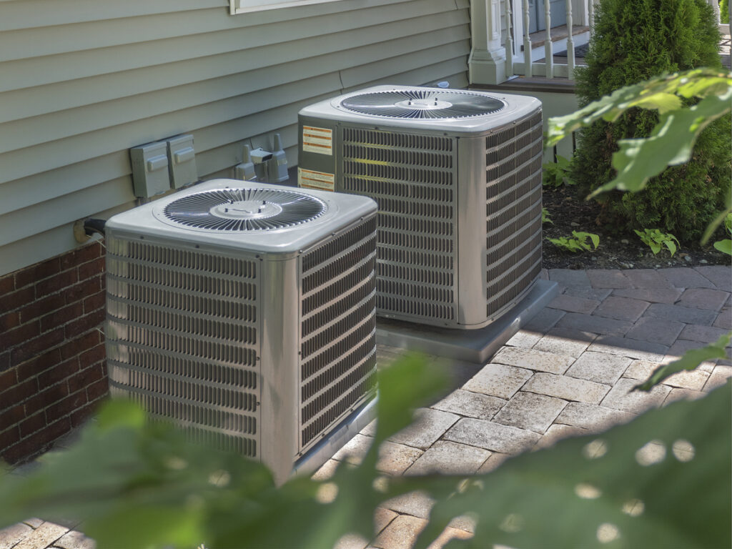 Two outdoor HVAC condenser units installed on a brick patio beside a house.