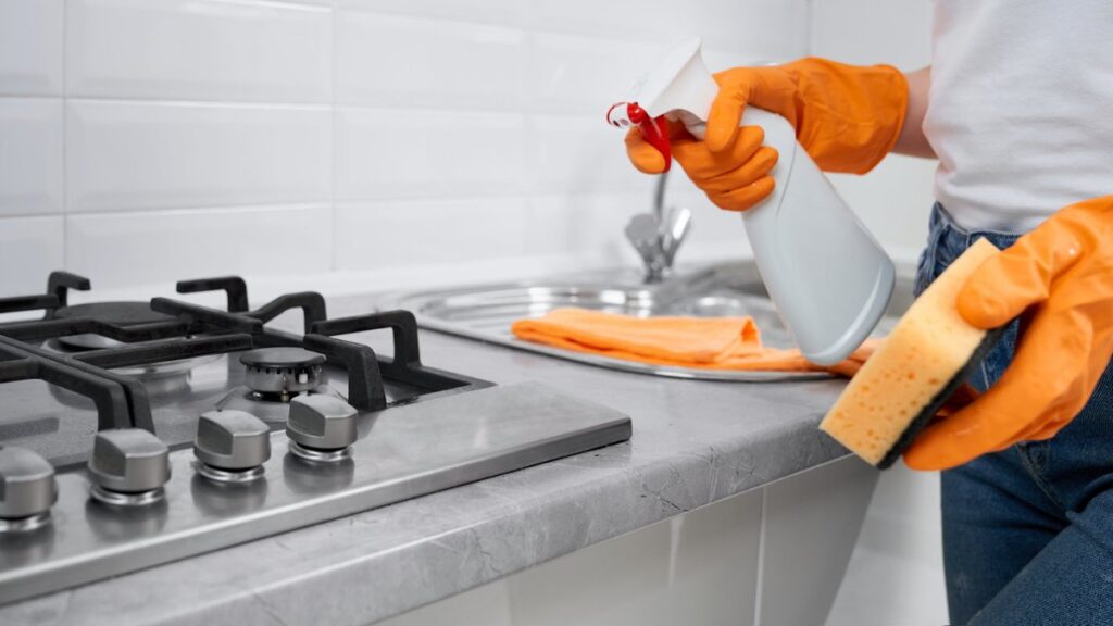 Person wearing orange gloves holding a spray bottle and a sponge to clean a kitchen countertop.