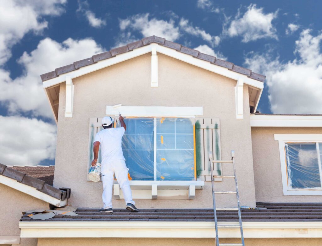 A house painter in white stands on a roof, painting the white trim of a second-story window.