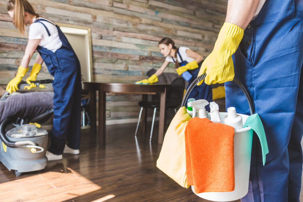 Professional cleaners in blue uniforms cleaning a room, with a bucket of supplies in the foreground.