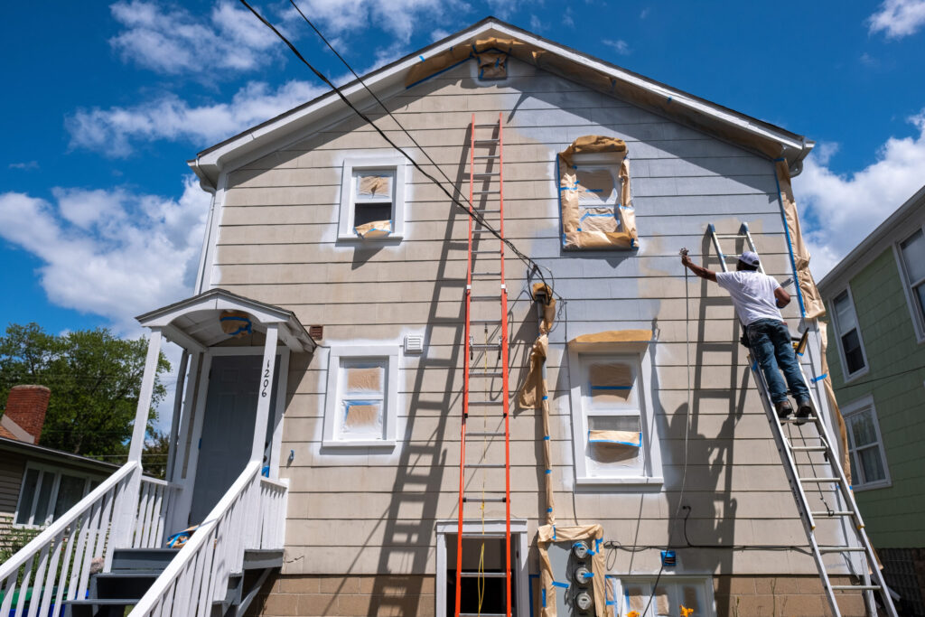 Painter on a ladder spray-painting the exterior wall of a two-story house with taped-off windows.