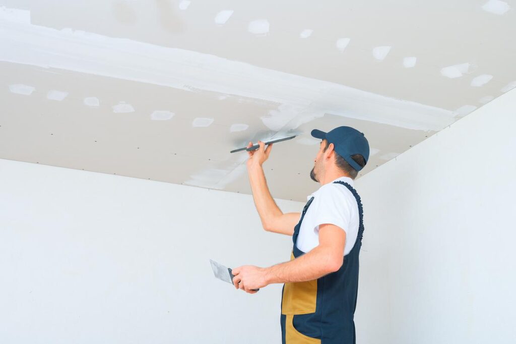 Construction worker applying plaster to a drywall ceiling with a trowel.