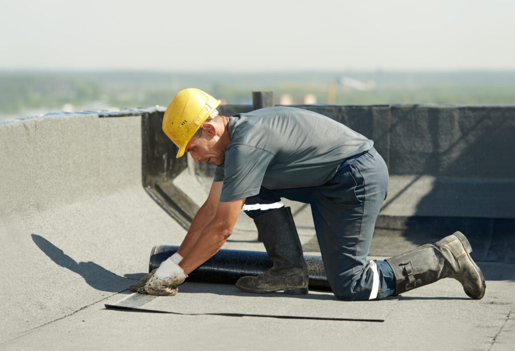 Worker in a yellow hard hat and protective gear installs waterproof roofing material on a flat roof.