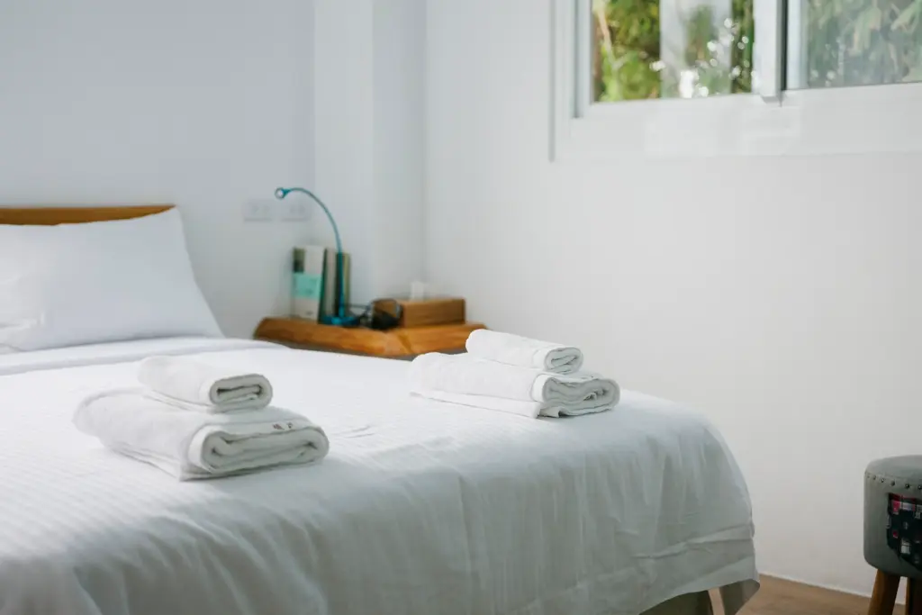 Neatly folded white towels stacked on a clean white bed in a bright minimalist hotel room.