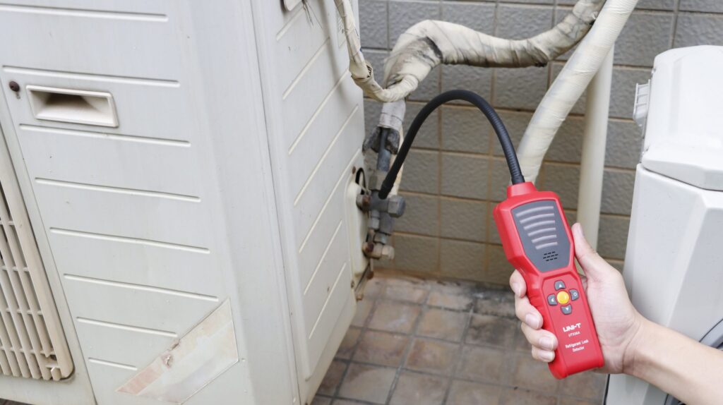 Hand using a red refrigerant leak detector on an outdoor air conditioning unit.