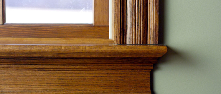 Close-up of a polished wood window sill and fluted trim against a sage green wall.