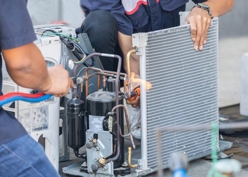 Technicians use a blowtorch to braze copper pipes on an air conditioning compressor and condenser unit.