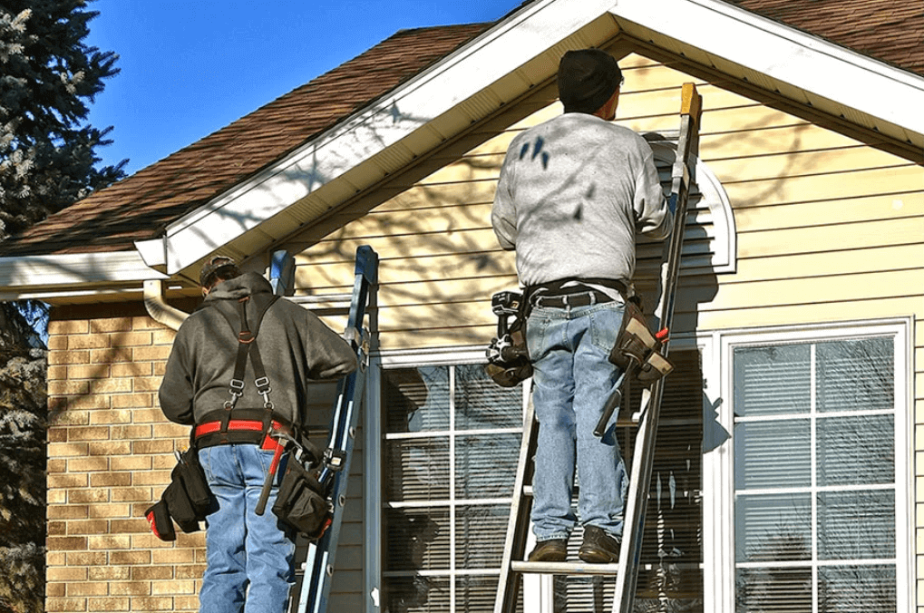 Two construction workers on ladders performing maintenance on the exterior of a house.
