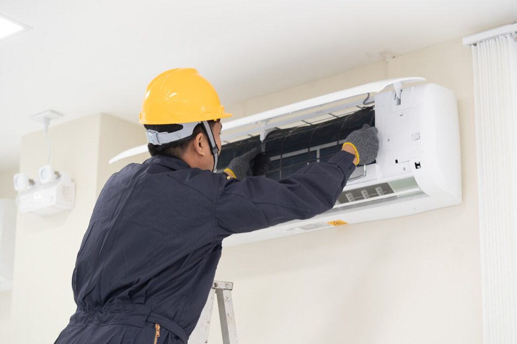 Technician in a yellow hard hat and blue jumpsuit servicing a wall-mounted air conditioner.