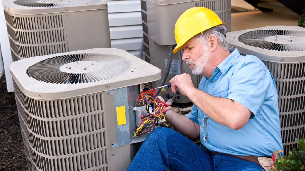 HVAC technician in a yellow hard hat repairing the wiring on an outdoor air conditioner unit.