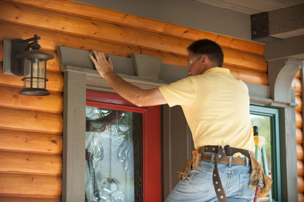 A worker in a yellow shirt on a ladder installs grey trim above a red door on a log cabin house.