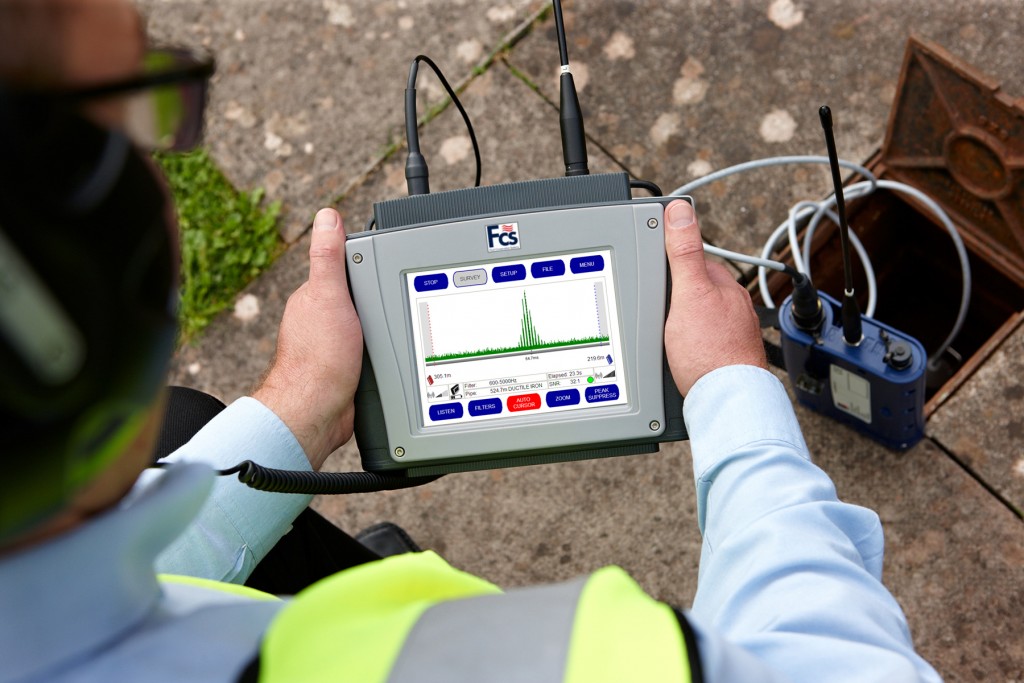 A worker in high-visibility gear holds a handheld diagnostic device displaying a frequency wave graph.