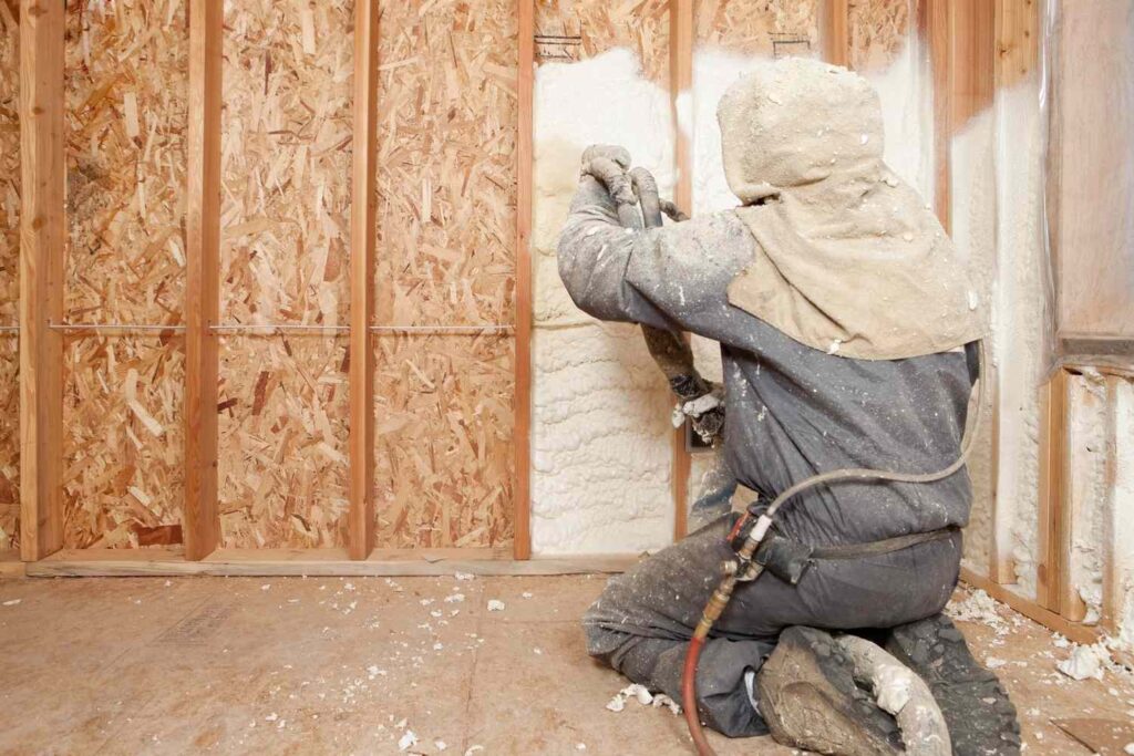 Worker in protective gear spraying foam insulation into wall studs of a home under construction.