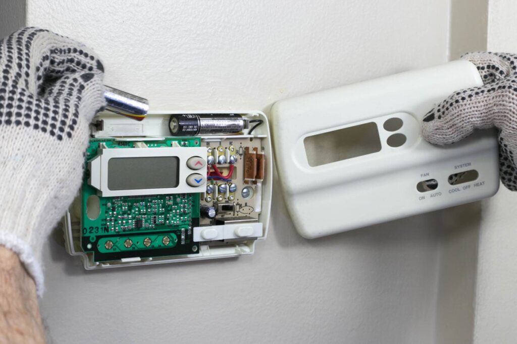 Gloved hands replacing a battery in an open digital thermostat mounted on a wall.