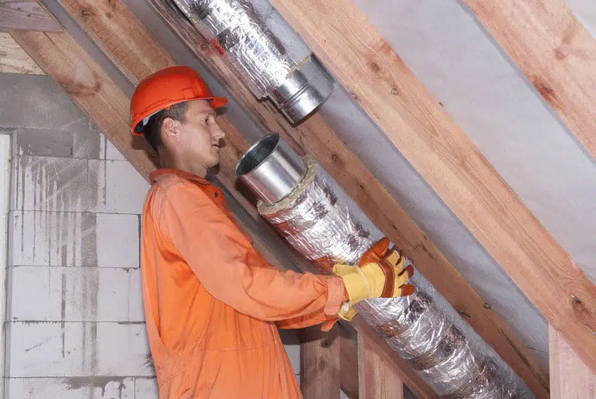 HVAC technician in orange coveralls and hard hat installing insulated ductwork in a wooden attic.