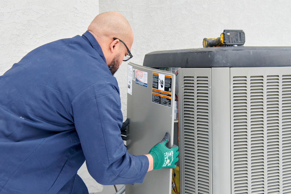 HVAC technician in a blue uniform servicing an outdoor AC condenser unit.