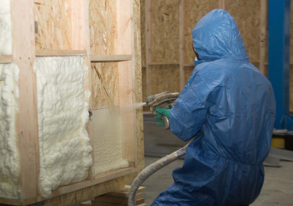 Worker in blue protective gear spraying white foam insulation into wooden wall studs.