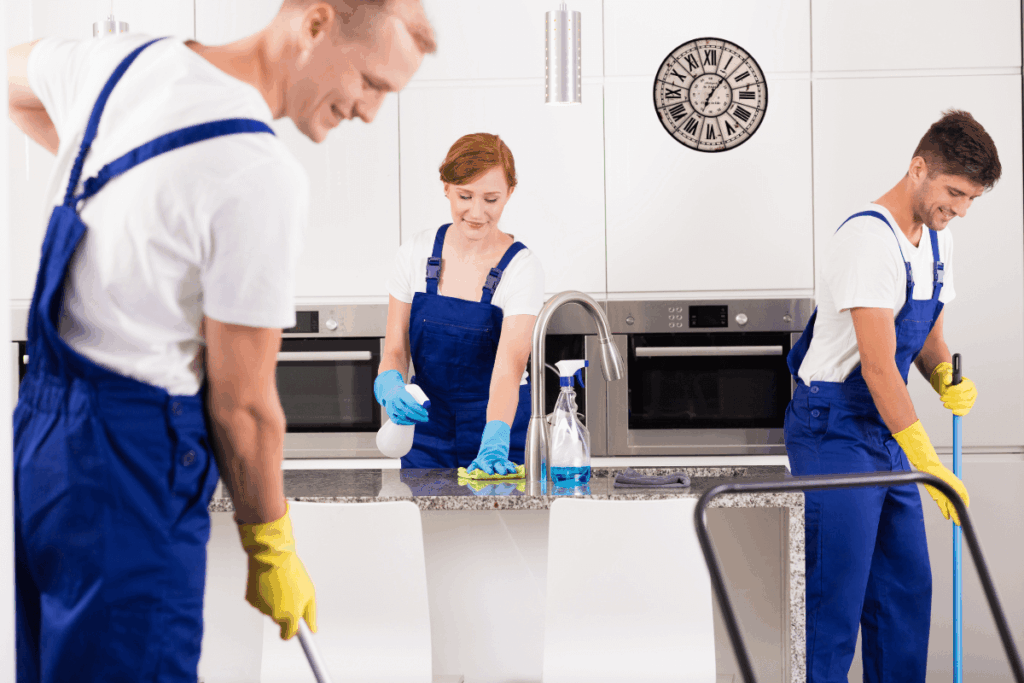 Three professional cleaners in blue uniforms and yellow gloves cleaning a modern kitchen.