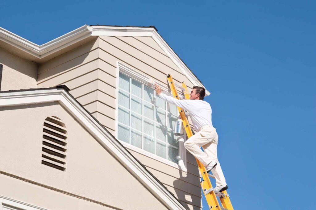 A man on a ladder paints the trim of a house against a clear blue sky.