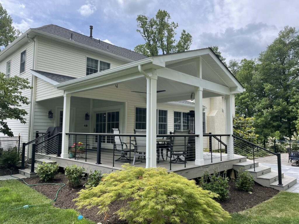 Covered white back deck with cable railing, gabled roof, and outdoor seating attached to a two-story house.