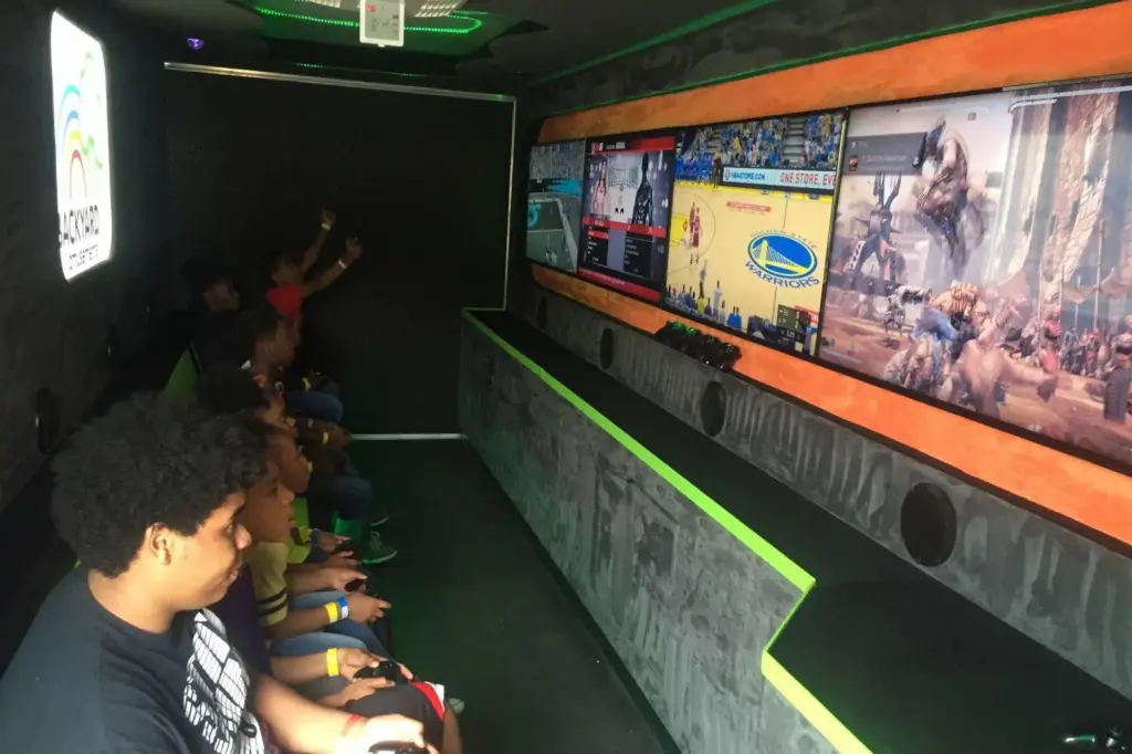 Children playing video games in a mobile gaming truck with multiple wall-mounted screens.