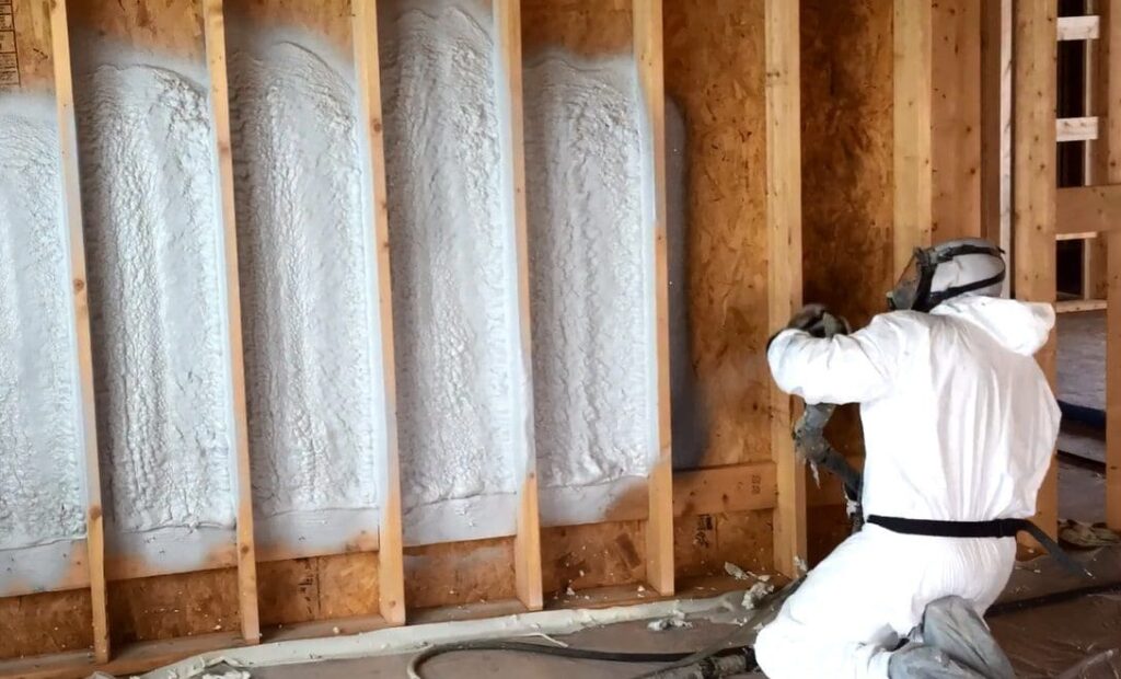Worker in protective gear applying spray foam insulation to wooden wall studs.