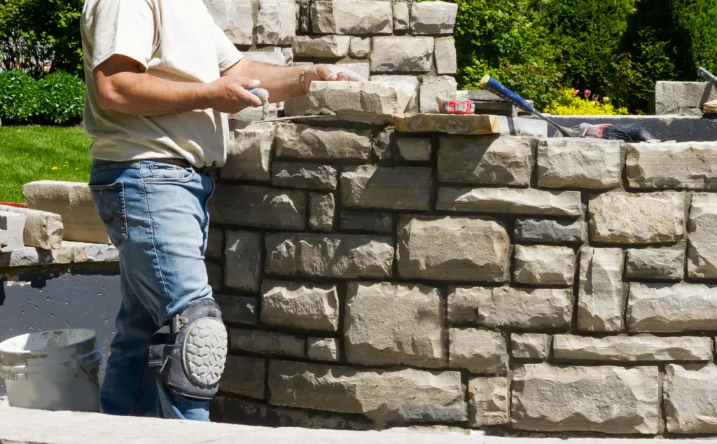 A stone mason wearing a knee pad applies mortar to build a natural stone wall in a garden setting.