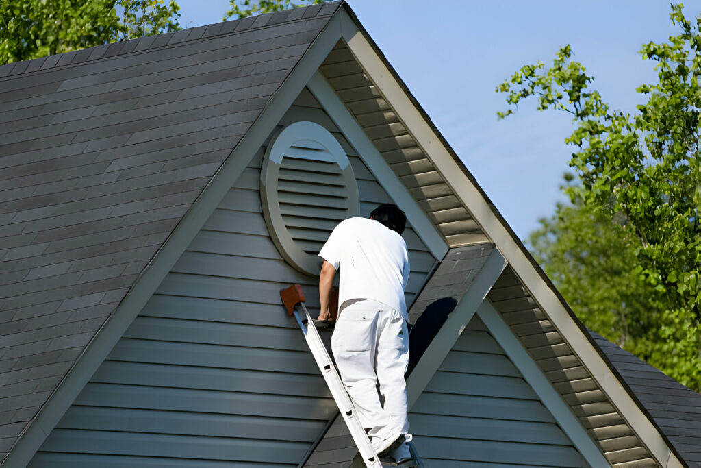 Person on a ladder painting a circular vent on the gable of a grey house.