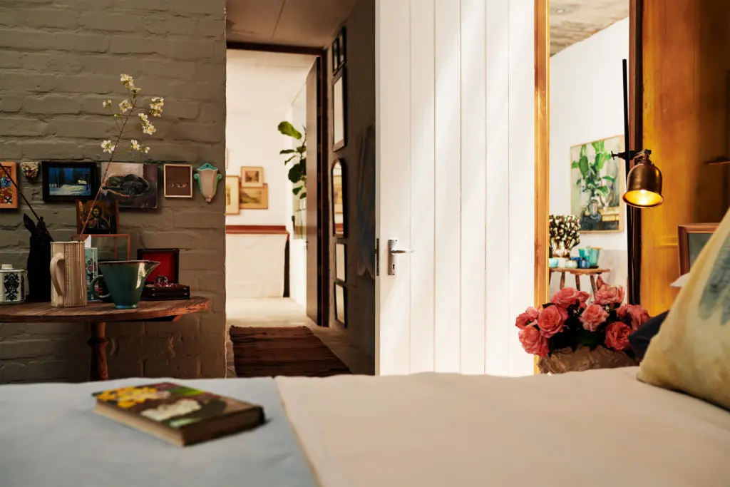 View through a bedroom into a hallway and adjacent room with eclectic decor, flowers, and warm lighting.