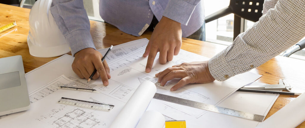 Architects collaborating on blueprints at a wooden table with rulers, pencils, and a hard hat.