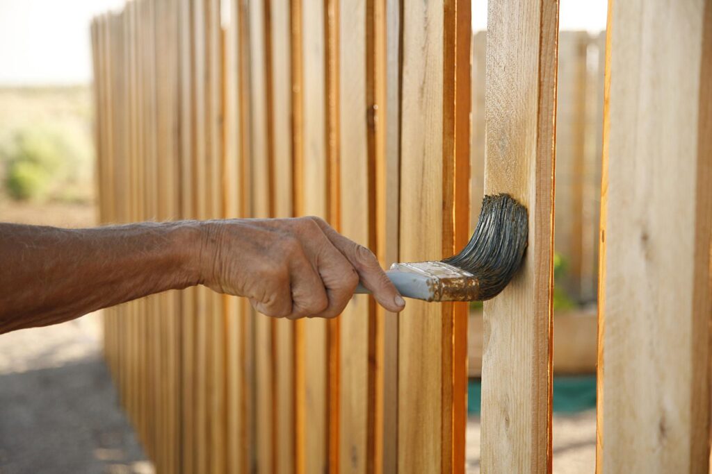 A hand using a paintbrush to apply brown stain to a wooden fence.