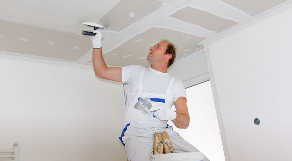 A plasterer in white overalls applying joint compound to a drywall ceiling with a trowel.