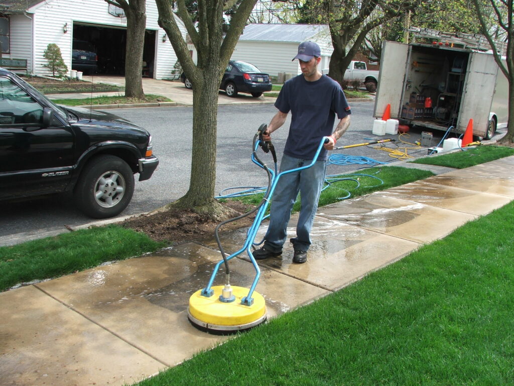 Man using a yellow power washing surface cleaner on a residential concrete sidewalk.