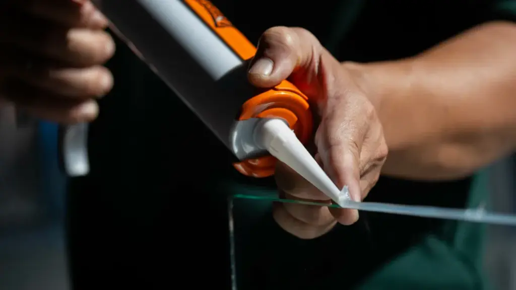 Close-up of a hand using a caulking gun to apply a thin bead of silicone sealant along the edge of a glass pane.