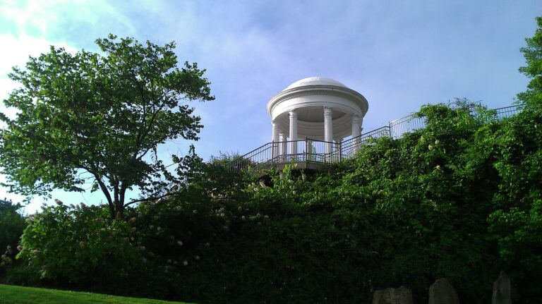 White domed gazebo with classical columns perched on a lush, green hillside under a clear blue sky.