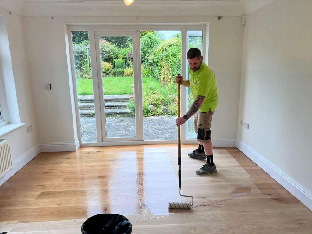 Man applying liquid sealant to a light wood floor using a roller on an extension pole.
