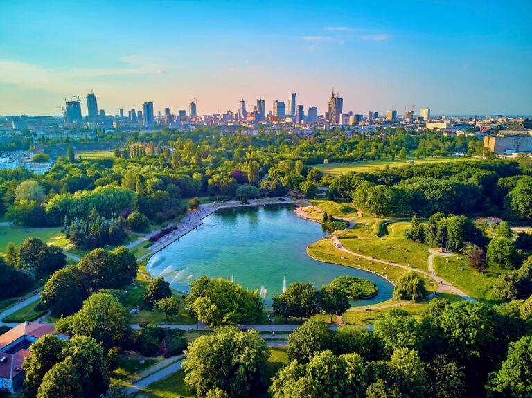 Aerial view of a lush city park with a central blue pond and a distant urban skyline.