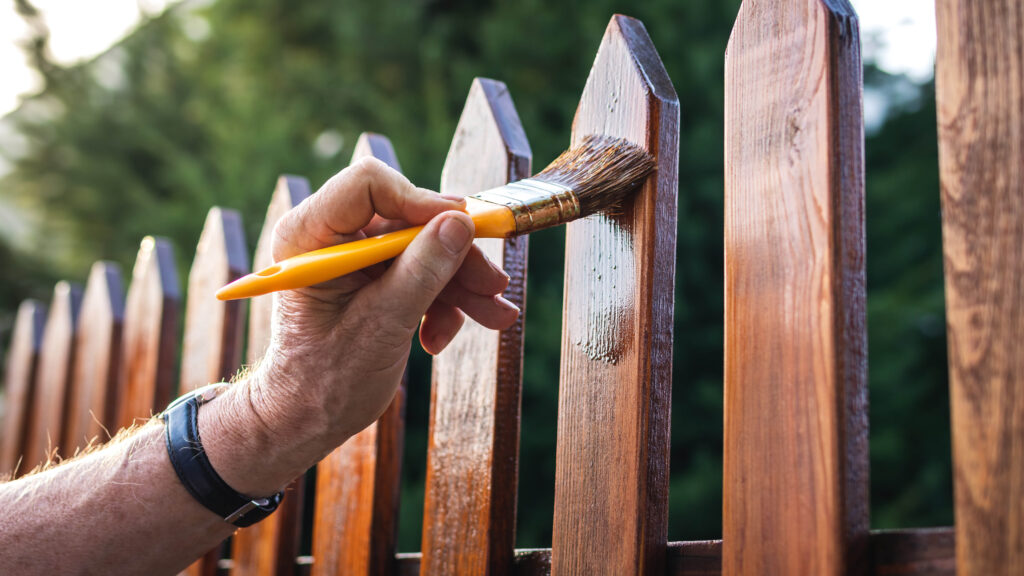 A person's hand using a yellow-handled brush to apply wood stain to a picket fence.