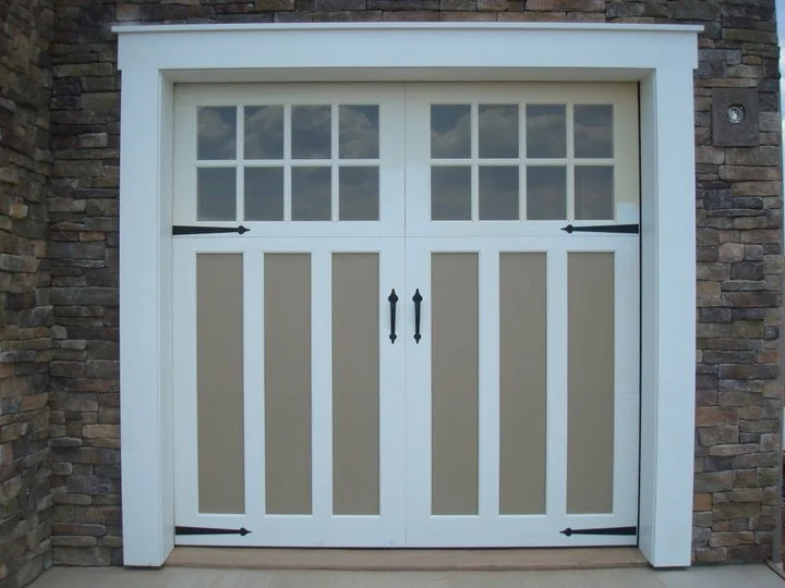 White and tan carriage-style garage door with window panes and black hardware, set against a stone wall.