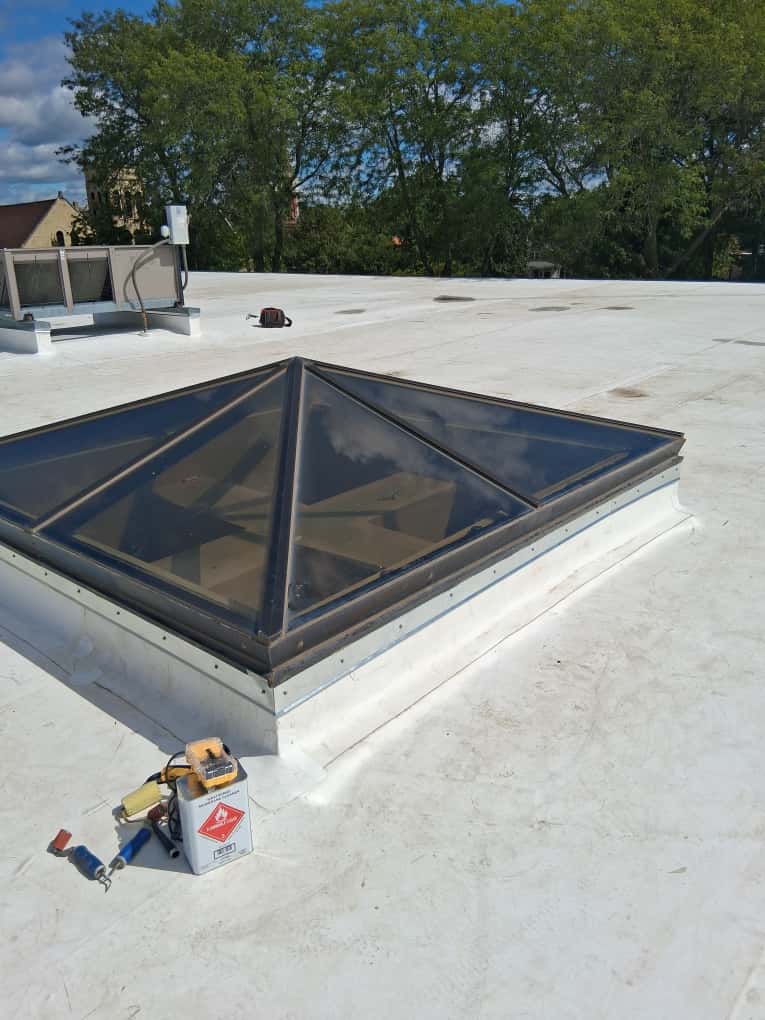 Pyramid skylight on a flat white roof with maintenance tools and sealant nearby.