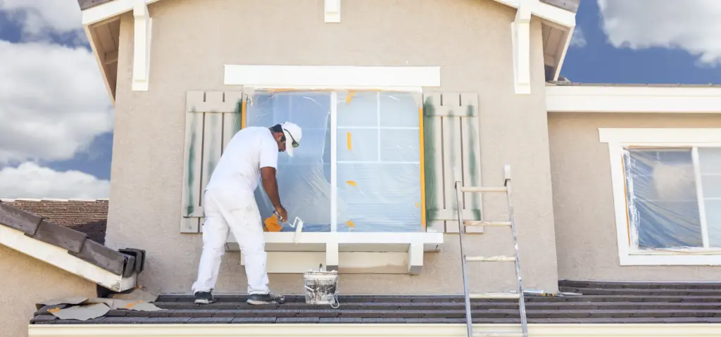 A painter in white clothing uses a roller to paint a window frame on the exterior of a house.