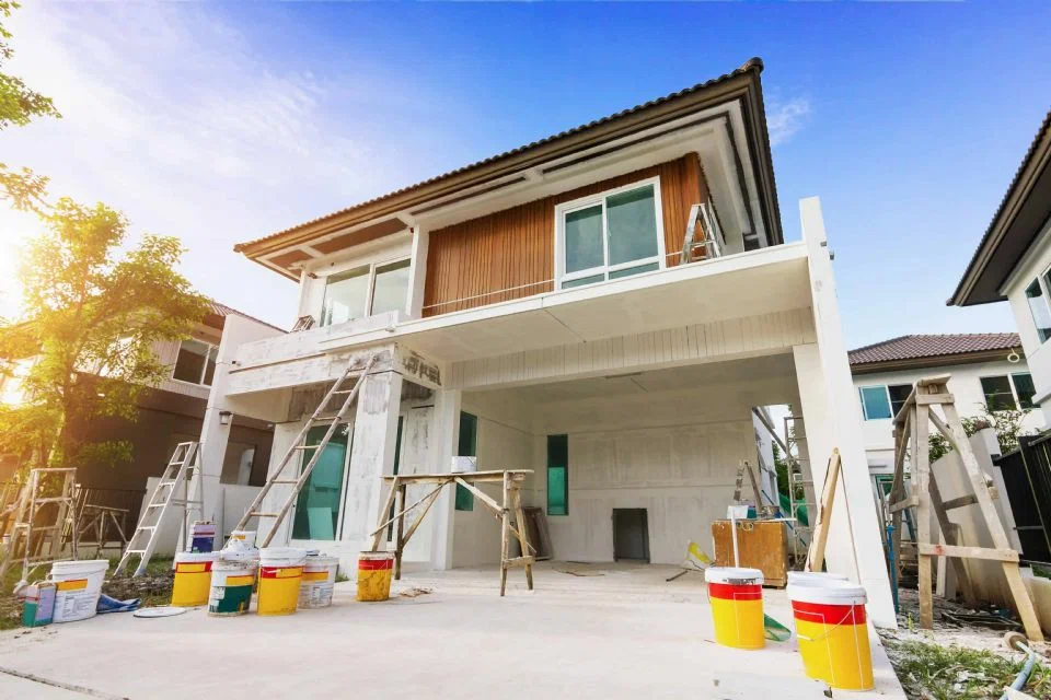 Modern house undergoing exterior renovation with ladders, scaffolding, and paint cans in the driveway.